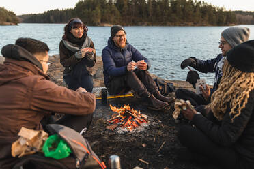 Group of smiling multiethnic sitting around campfire on coast of pond and playing cards - ADSF26403