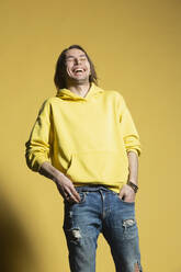 Glücklicher Mann in Jeans und Kapuzenpulli lachend vor gelbem Hintergrund - FSIF05698