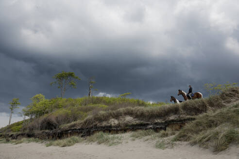 Mädchen auf dem Pferderücken im Strandgras unter stürmischem Himmel - FSIF05703
