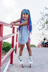 Fröhliches ethnisches Kind mit herausgestreckter Zunge und bunten Zöpfen beim Rollschuhlaufen und Blick in die Kamera in der Stadt - ADSF26417
