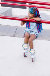 Cheerful Mexican kid with bright braids in roller skates sitting while leaning with hands on fence and looking away - ADSF26418