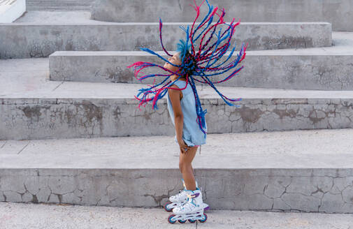 Side view of ethnic child in roller skates with flying colorful braids standing on staircase - ADSF26419