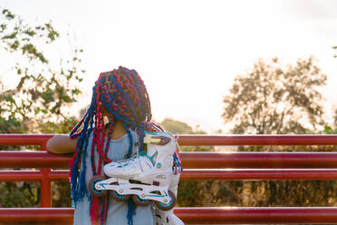 Back view of anonymous Mexican kid with colorful braids admiring trees while leaning on fence in evening - ADSF26422