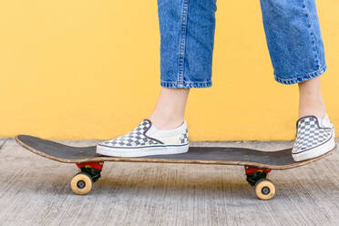 Cropped unerkennbar weiblichen Skater mit Skateboard stehen auf Gehweg mit bunten gelben Wand auf dem Hintergrund in der Tageszeit - ADSF26433