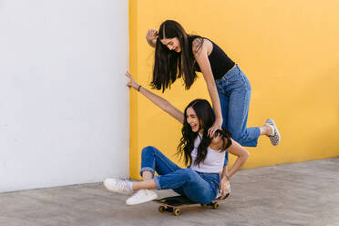 Cheerful teen with crossed legs riding skateboard with content female sibling on walkway in daytime - ADSF26434