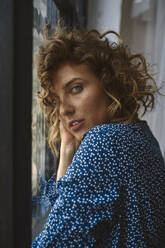 Side view of charming curly woman touching face and thoughtfully looking at camera near windows - ADSF26465