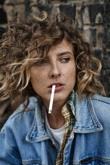 Stylish curly woman sitting and thoughtfully smoking in abandoned place looking away - ADSF26466