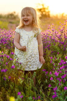 Adorable kleines Mädchen im weißen Kleid lachend und wegschauen, während stehend inmitten von schönen Blumen in erstaunlichen Wiese - ADSF26468