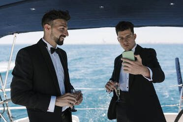 Smiling man looking at male friend taking selfie in yacht during vacation - EGAF02436