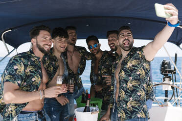 Mann streckt Zunge heraus, während er ein Selfie mit männlichen Freunden während einer Party auf einer Jacht macht - EGAF02446