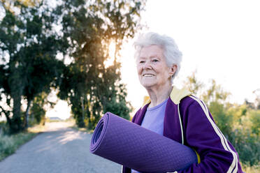 Optimistische ältere Frau trägt eine zusammengerollte Matte und lächelt vor dem Fitnesstraining am Morgen auf dem Lande - ADSF26479