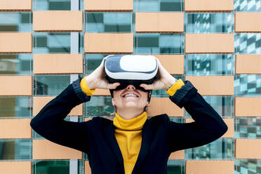 Smiling female in jacket and turtleneck using VR goggles near building windows in daylight - ADSF26480