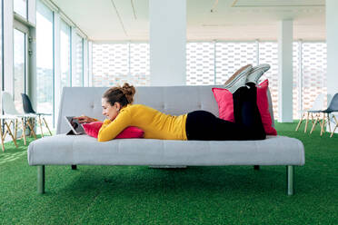 Positive adult female in casual clothes lying on couch with pillow while browsing on tablet in light living room - ADSF26483