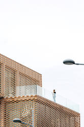 Von unten der weiblichen in stilvollem Outfit stehen auf dem Balkon des modernen Gebäudes mit geometrischen Elementen auf Fenster, während mit Tablet in der Nähe von Glas-Geländer unter hellem Himmel - ADSF26489