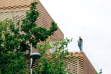 Von unten der weiblichen in stilvollem Outfit stehen auf dem Balkon des modernen Gebäudes mit geometrischen Elementen auf Fenster, während mit Tablet in der Nähe von Glas Geländer unter hellem Himmel in der Nähe von grünen Bäumen - ADSF26490