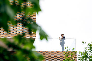 Von unten der weiblichen in stilvollem Outfit stehen auf dem Balkon des modernen Gebäudes mit geometrischen Elementen auf Fenster, während mit Tablet in der Nähe von Glas Geländer unter hellem Himmel in der Nähe von grünen Bäumen - ADSF26491