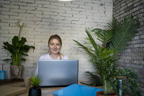Smiling young female professional using laptop at table in home office - RCPF01179
