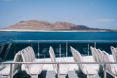 Empty white chairs on deck of cruise boat sailing in blue sea water with mountain on shore - ADSF26532