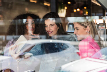 Through glass of cheerful young multiracial female best friends in trendy outfits smiling and showing two fingers gesture while taking selfie on smartphone standing near window - ADSF26545