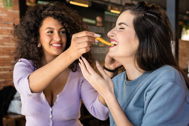 Positive junge ethnische Frau mit dunklen lockigen Haaren füttert hungrige fröhliche Freundin mit appetitlichen Pommes frites im Restaurant - ADSF26546