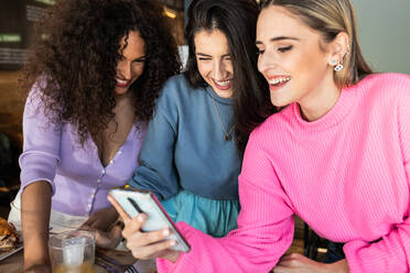 Lächelnde junge Freundinnen in Freizeitkleidung, die beim Mittagessen im Restaurant auf ihrem Handy surfen - ADSF26547