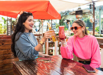 Glückliche junge Freundinnen in legerer Kleidung stoßen mit Bierflaschen und Beerencocktails an, während sie an einem Tisch in einer sonnigen Bar im Freien sitzen - ADSF26550