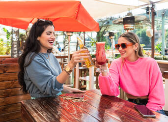 Happy young female friends in casual outfits clinking beer bottle and glass of berry cocktail while sitting at table in sunny outdoor bar - ADSF26550