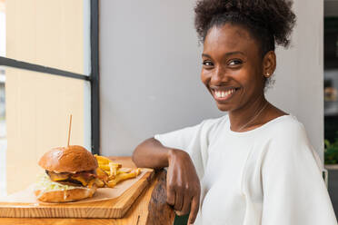 Afroamerikanische Frau isst leckere Pommes frites und leckere Hamburger, die auf einem Holzbrett auf einem hohen Tisch in einem Schnellrestaurant serviert werden, während sie in die Kamera schaut - ADSF26554
