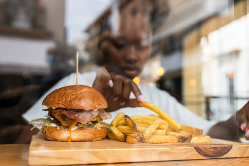 Durch die Glaswand verschwommene afroamerikanische Frau isst leckere Pommes frites und leckere Hamburger, die auf einem Holzbrett auf einem hohen Tisch in einem Schnellrestaurant serviert werden - ADSF26555