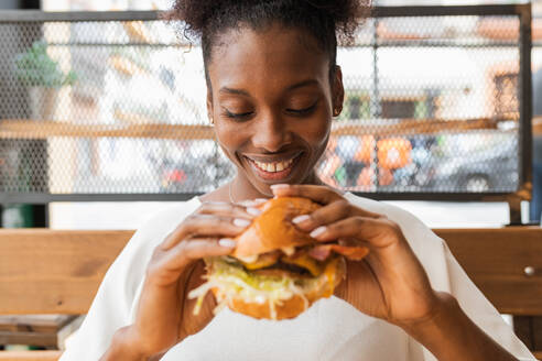 Crop African American weiblich in Freizeitkleidung Essen leckeren frischen Cheeseburger in hellen Raum - ADSF26557