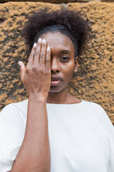 Emotionless young African American female in white shirt covering half face with hand and looking at camera against uneven rough wall - ADSF26559