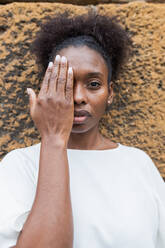 Emotionslose junge Afroamerikanerin in weißem Hemd, die ihr halbes Gesicht mit der Hand bedeckt und in die Kamera gegen eine unebene, raue Wand schaut - ADSF26559