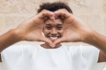 Glückliche junge Afroamerikanerin in weißer Bluse, die mit den Händen ein Herzzeichen zeigt und mit einem breiten Grinsen in die Kamera schaut, während sie an einer unebenen Wand steht - ADSF26560