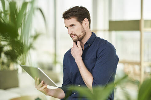 Thoughtful businessman with hand on chin holding digital tablet at home - AKLF00318