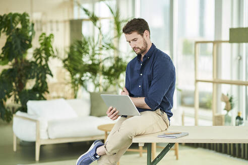 Männlicher Fachmann, der auf ein digitales Tablet schaut, während er auf einem Tisch im Heimbüro sitzt - AKLF00321