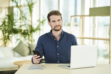 Businessman holding mobile phone while sitting with laptop in home office - AKLF00327