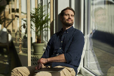 Smiling male professional with eyes closed leaning on glass window at home - AKLF00328