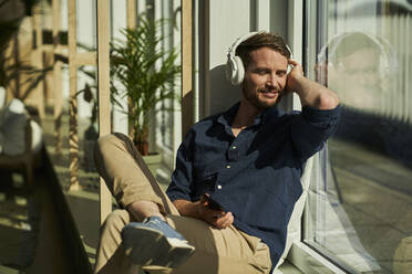 Smiling male freelancer with eyes closed listening music through wireless headphones by window - AKLF00329