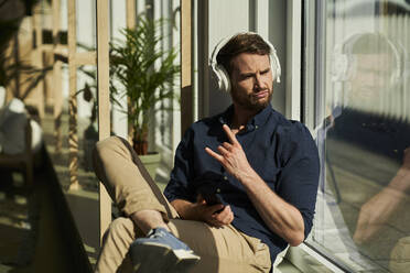 Businessman gesturing horn sign while listening music through wireless headphones by window - AKLF00330