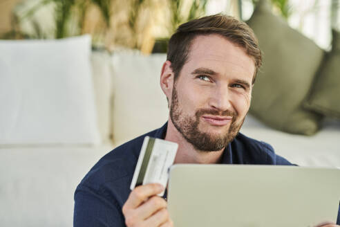 Männlicher Freiberufler, der die Lippen spitzt, während er eine Kreditkarte zu Hause hält - AKLF00338