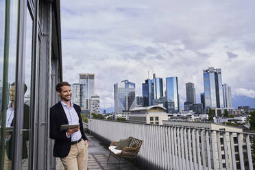Lächelnder männlicher Fachmann, der ein digitales Tablet hält, während er mit der Hand in der Tasche auf einer Gebäudeterrasse steht - AKLF00343