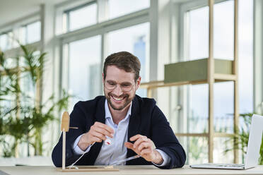 Glücklicher männlicher Freiberufler mit Brille, der ein architektonisches Modell im Heimbüro anfertigt - AKLF00353