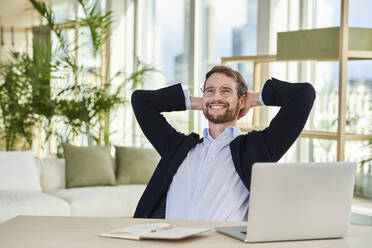 Happy relaxed male freelancer sitting with hands behind head in home office - AKLF00359