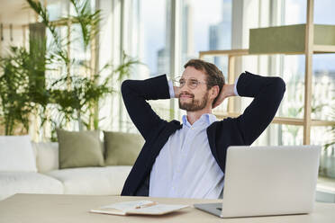 Smiling relaxed businessman sitting with hands behind head in home office - AKLF00360