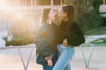 Smiling diverse lesbian girlfriends in trendy wear embracing and speaking while looking at each other on walkway - ADSF26566