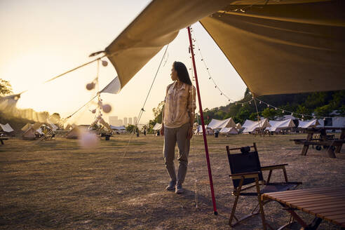 Ethnische asiatische Frau mit Sonnenbrille, die sich beim Sonnenuntergang auf einem Campingplatz entspannt und wegschaut - ADSF26572