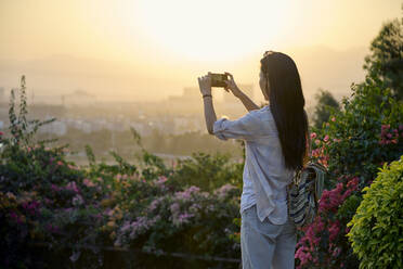 Ethnische asiatische Frau steht im Garten und fotografiert mit ihrem Smartphone bei Sonnenuntergang - ADSF26573
