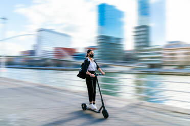 Ethnic female entrepreneur in fabric mask and formal clothes riding electric scooter on walkway in town during coronavirus pandemic - ADSF26576