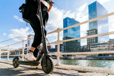 Seitenansicht der Ernte anonyme Frau in Turnschuhen Reiten modernen Roller auf dem Gehweg gegen Stadt Fluss - ADSF26577