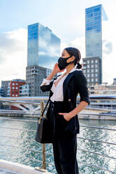 Ethnische Unternehmerin in formeller Kleidung und Gesichtsmaske aus Stoff mit der Hand in der Tasche, die mit dem Handy spricht, während sie in der Stadt wegschaut - ADSF26583